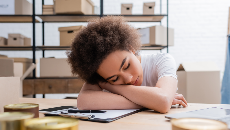 Zmęczona młoda kobieta śpiąca przy biurku w biurze, oparta głową na rękach, wokół niej kartony i dokumenty.