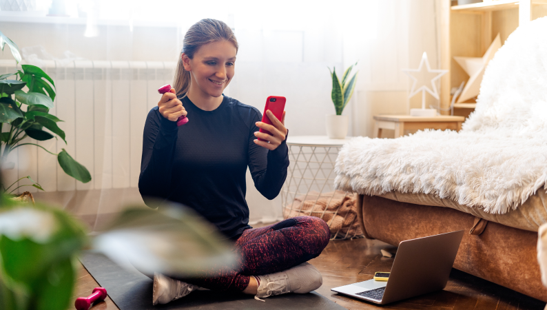 Kobieta siedzi na macie do ćwiczeń z telefonem i hantlami, uśmiechnięta podczas aktywnego poranka – przykład na detoks cyfrowy w domu.