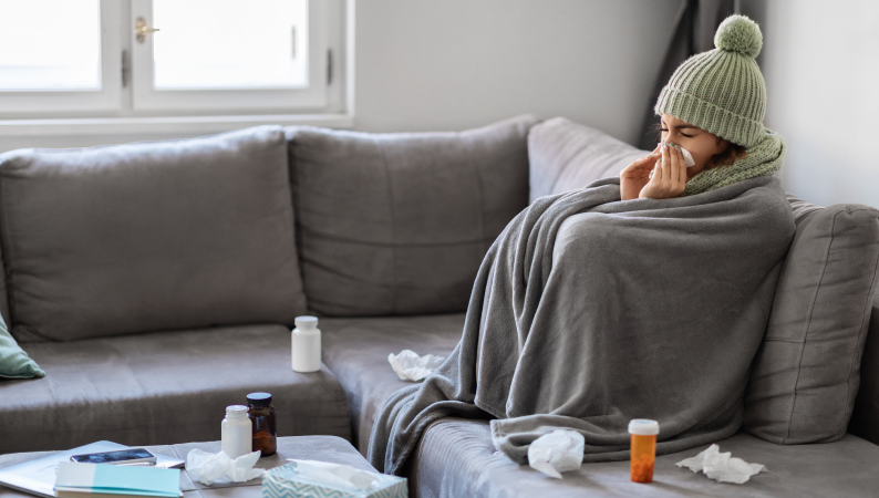 Kobieta otulona kocem i w czapce siedzi chora na kanapie – ilustracja potrzeby budowania odporności przez cały rok.