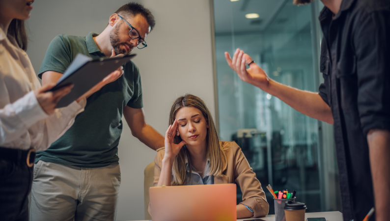 Kobieta przy biurku wygląda na zestresowaną, otoczona przez współpracowników – ilustracja potrzeby wsparcia, jakie dają adaptogeny na stres.