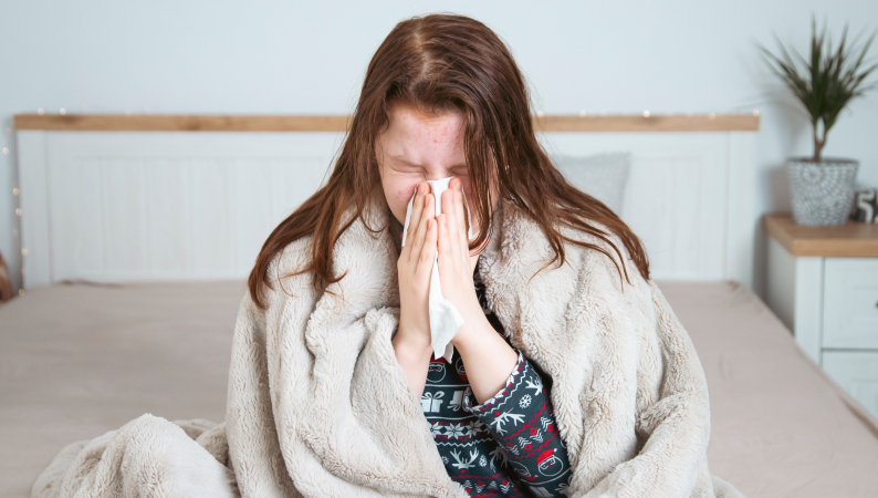 Kobieta chora na przeziębienie siedząca w łóżku pod kocem i wycierająca nos chusteczką – Reishi w sezonie przeziębień.
