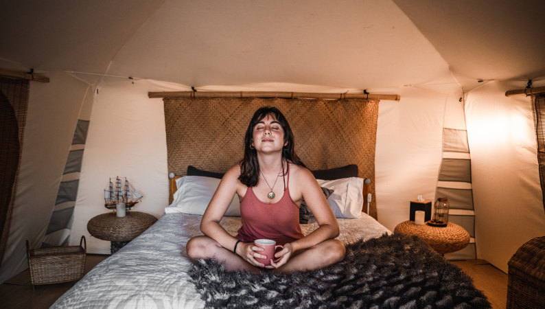 Uśmiechnięta kobieta siedząca po turecku na łóżku z kubkiem w dłoniach, relaksująca się podczas wieczornego rytuału na dobry sen.