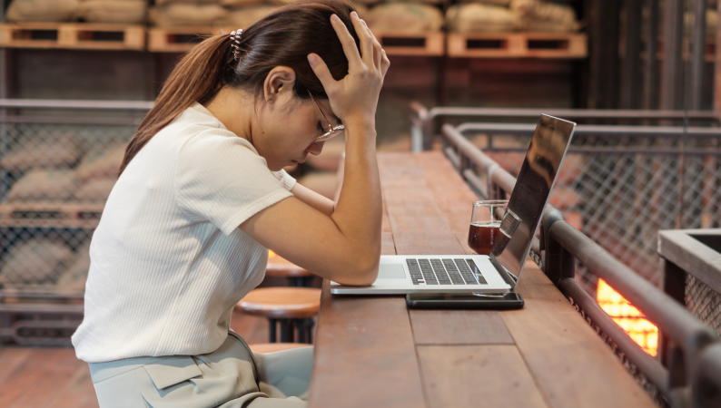 Kobieta przy laptopie trzymająca głowę w dłoniach – stres w pracy zdalnej i przemęczenie psychiczne spowodowane nadmiarem obowiązków.