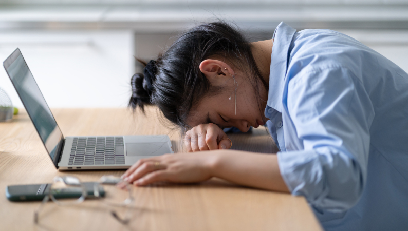 Kobieta opierająca głowę na biurku obok otwartego laptopa, wyczerpana pracą online – przykład objawu zjawiska cyfrowe zmęczenie.