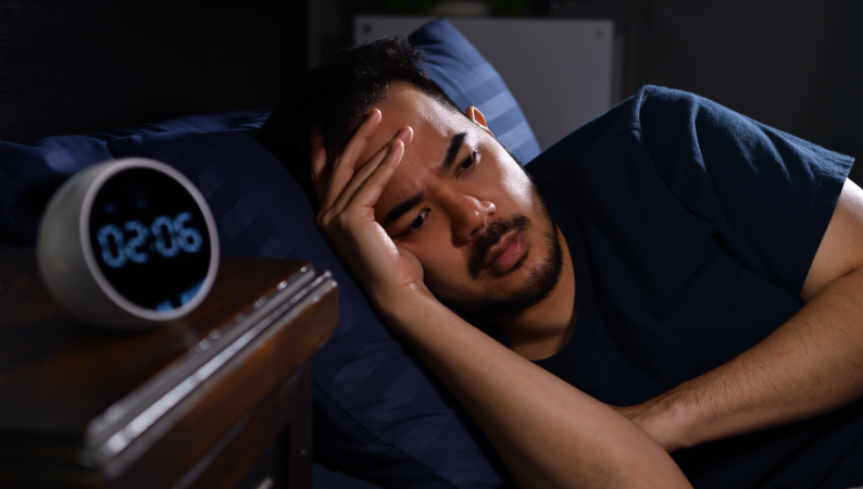 Mężczyzna leży w łóżku i patrzy na zegar, pokazując typowy problem nie możesz zasnąć mimo zmęczenia